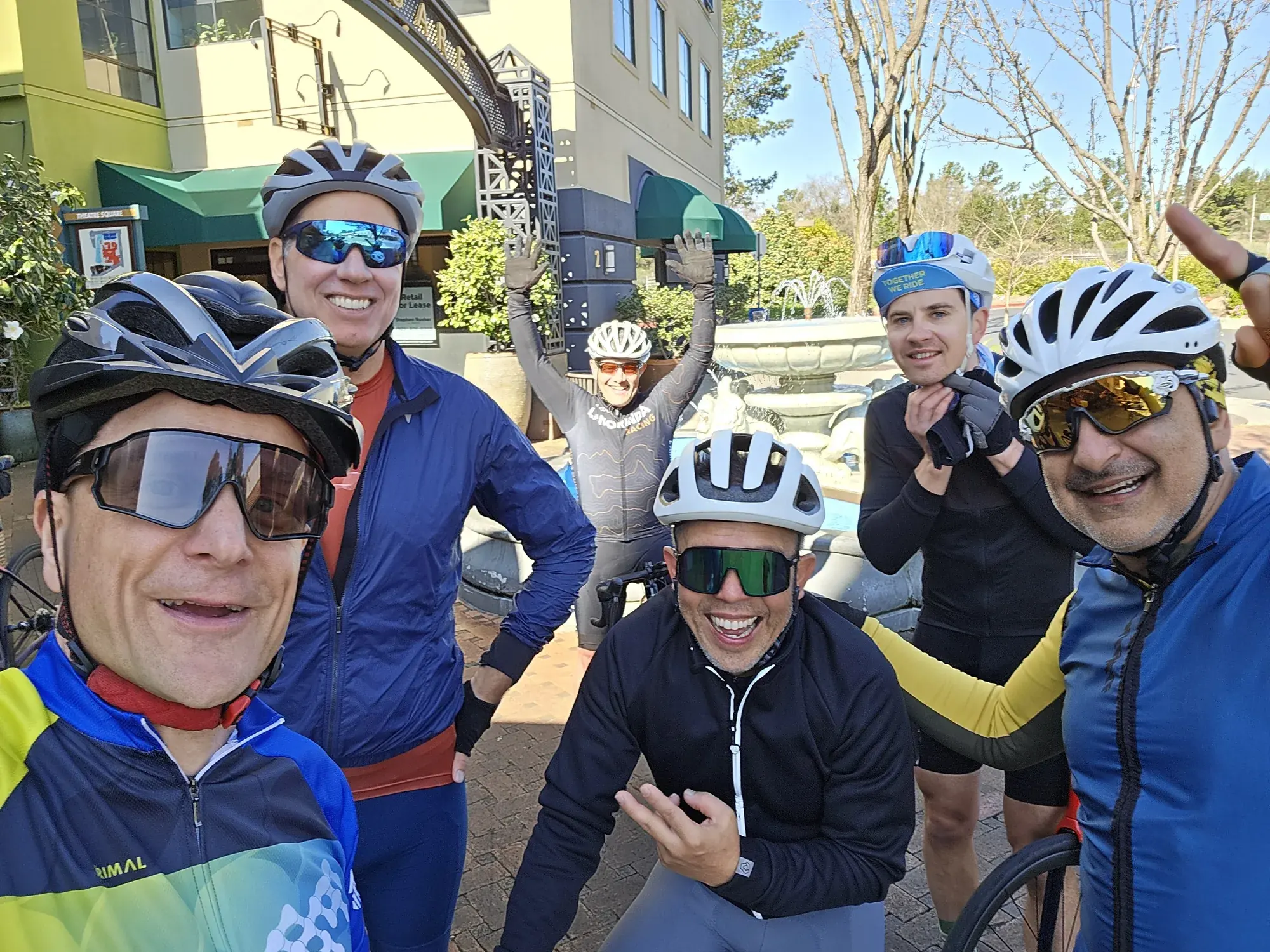 Six smiling cyclists gathered by the fountain at Theatre Square in Orinda